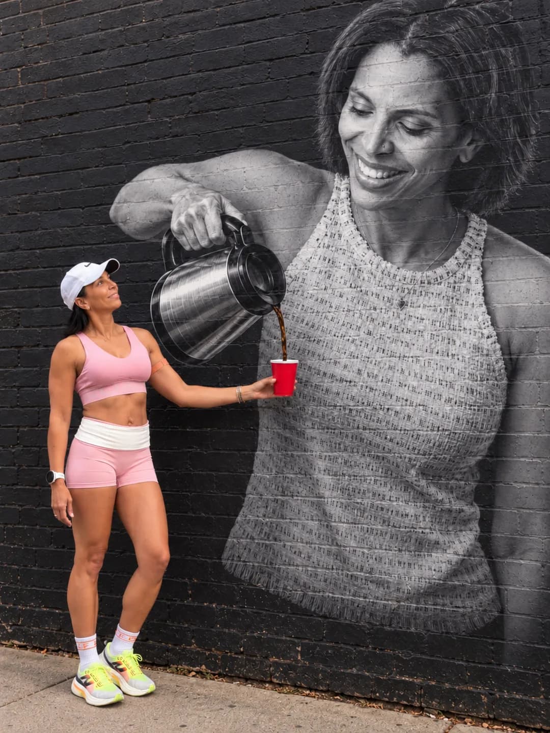 A woman in pink sportswear pours coffee into a cup from a giant black-and-white mural of herself holding a pitcher